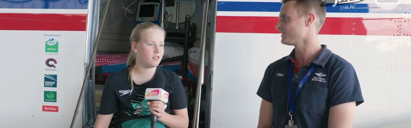 11 year old, Eliza, is pictured next to pilot, Elliot Johnston who is being interviewed. They are seated in front of an RFDS aeroplane, smiling.
