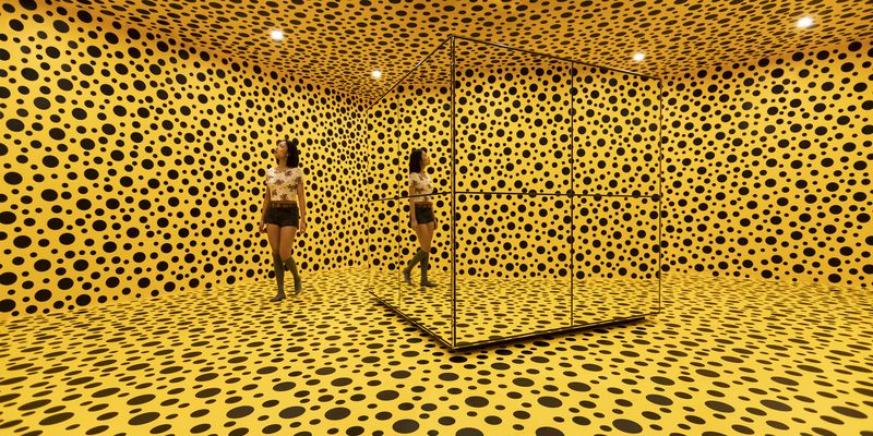 A young woman walks through bright yellow room with black circles on all four walls, ceiling and floor. There is a cube mirror at the centre of the room.