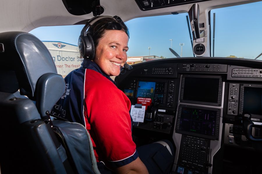 A lady in her early 30's stands next to a RFDS aircraft. She is wearing a red and navy blue RFDS operational uniform - shirt and pants. She is smiling