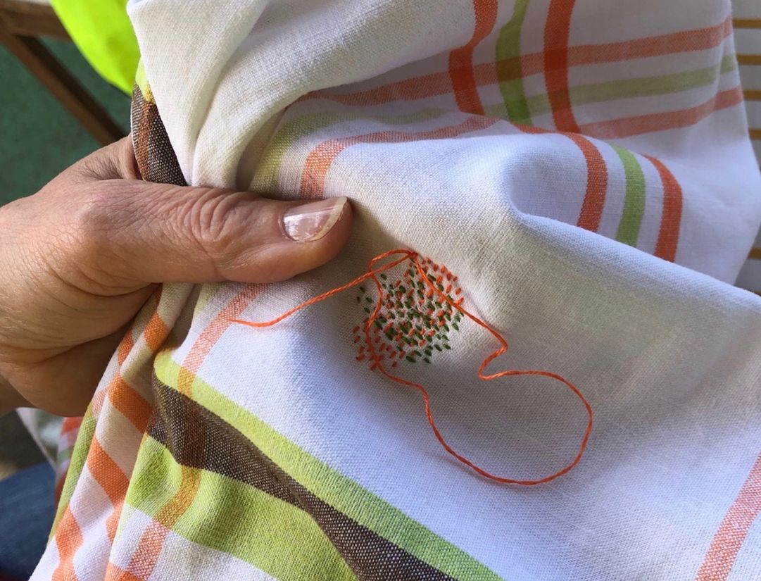 A close up of a hand with a needle mending a white tabecloth with green, brown and orange strips and orange thread