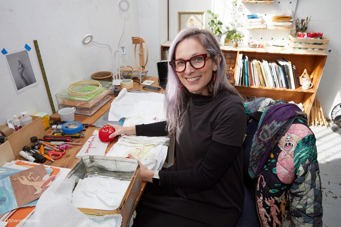 Photo of Sue Jo Wright at her desk in her studio with some textile work in progress pieces on the desk