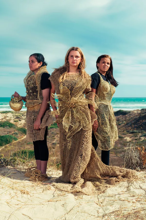 Three women stand in a coastal landscape wearing woven pieces made by First Nations artist Aunty Ellen Trevorrow