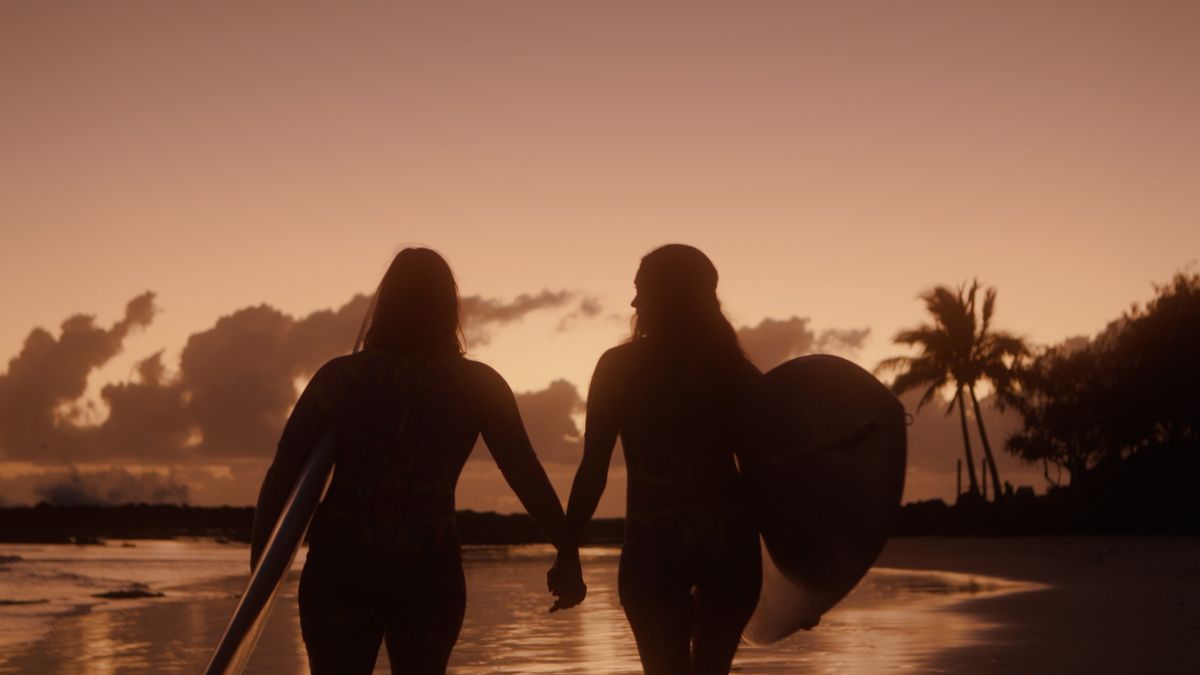 An image of two women in silhouette against a sunset walking with surfboards on the beach.