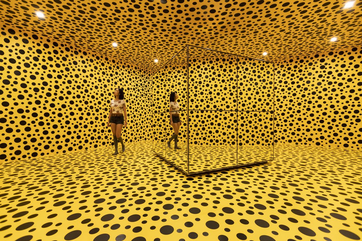 A young woman walks through bright yellow room with black circles on all four walls, ceiling and floor. There is a cube mirror at the centre of the room.