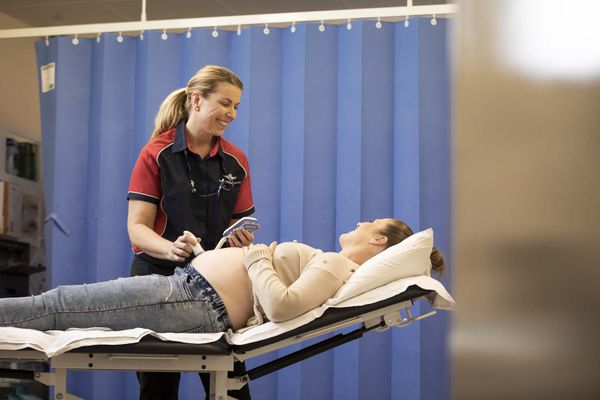 A pregnant woman lies on a hospital bed. A smiling female in a RFDS polo shirt holds a doppler over her pregnant tummy. They are both smiling. 