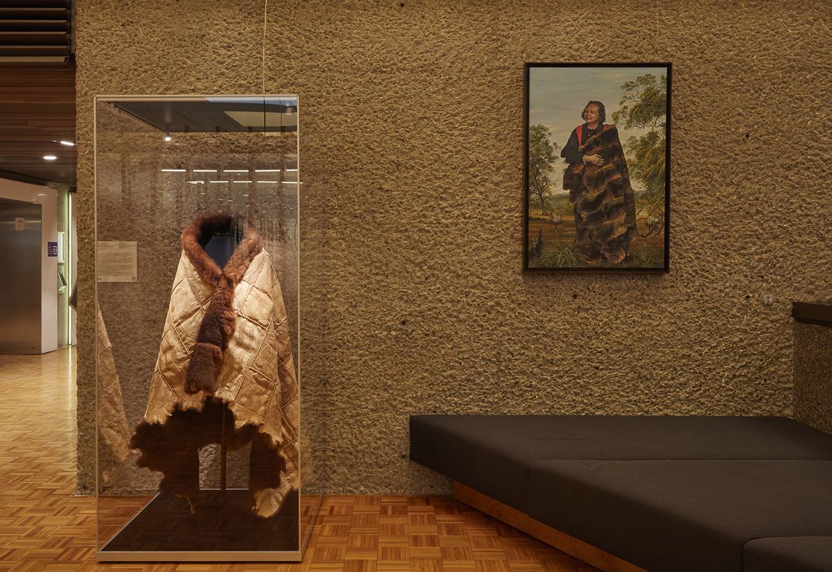 A photograph depicting a possum skin cloak in a museum case, next to a painted portrait of Aunty Joan Tranter wearing the same cloak.