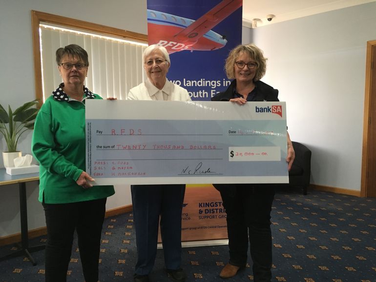 Three ladies smile at the camera. They are holding a novelty oversized cheque made out to the RFDS for $20,000. One lady is wearing a RFDS uniform. 