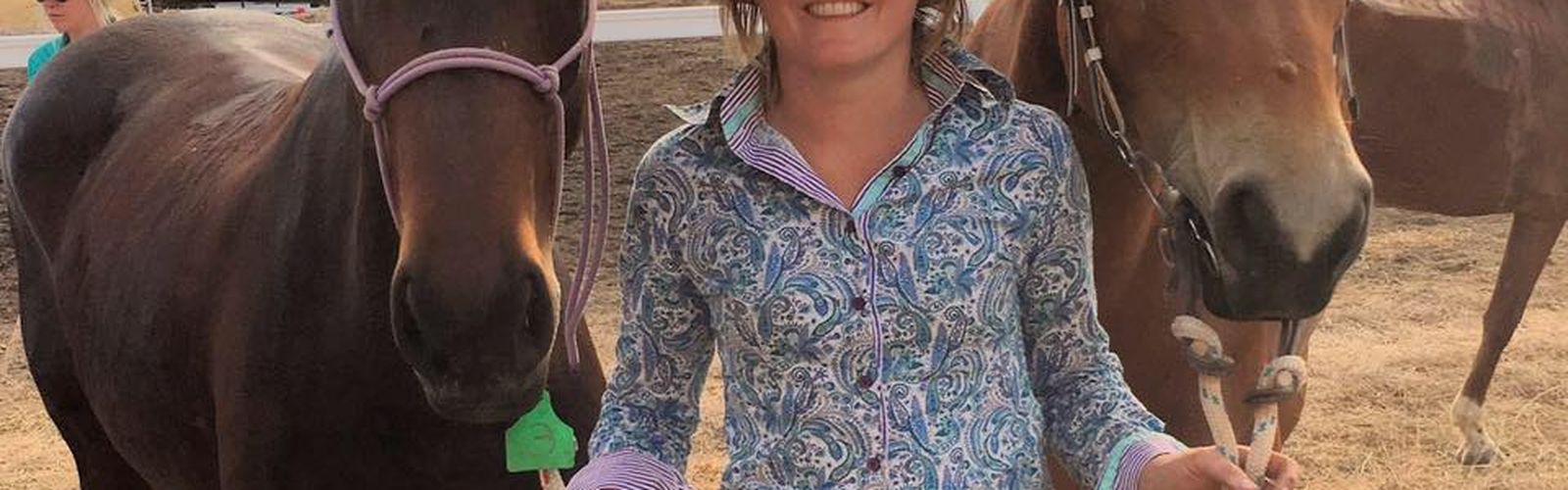 A young woman smiles. She is standing next to two horses. 