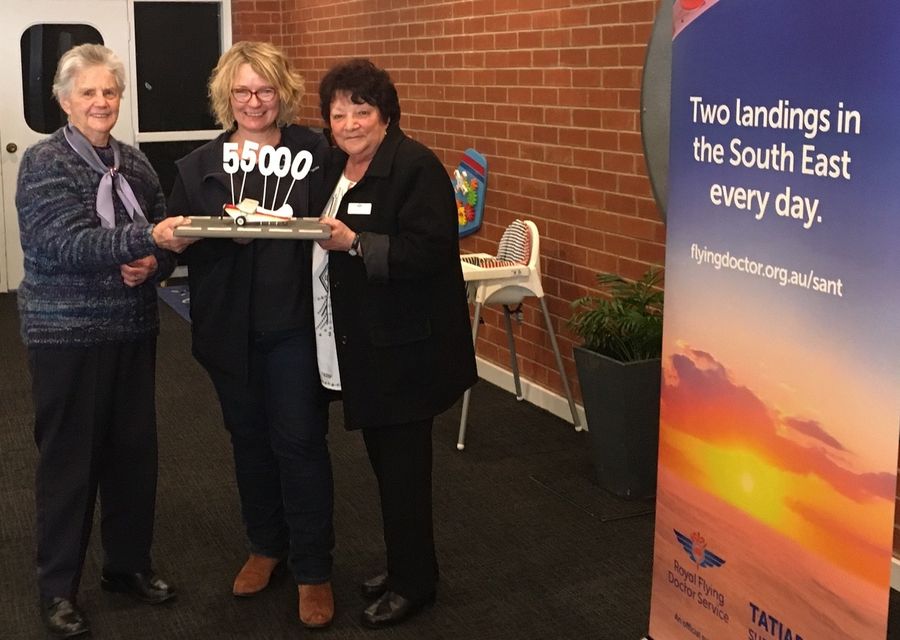 3 people holding a model plane smile at the camera. Figures spelling out 55000 are stuck onto the plane. 