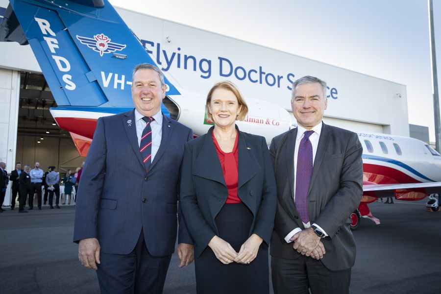 A woman and two men stand wearing business suits in front of a jet aircraft. The aircraft has a RFDS and S Kidman and Co logo on the side. 