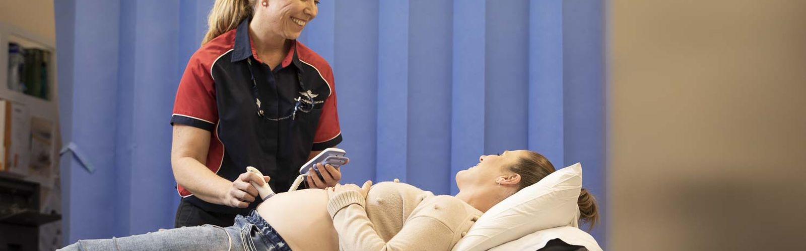 A pregnant woman lies on a hospital bed. A smiling female in a RFDS polo shirt holds a doppler over her pregnant tummy. They are both smiling. 