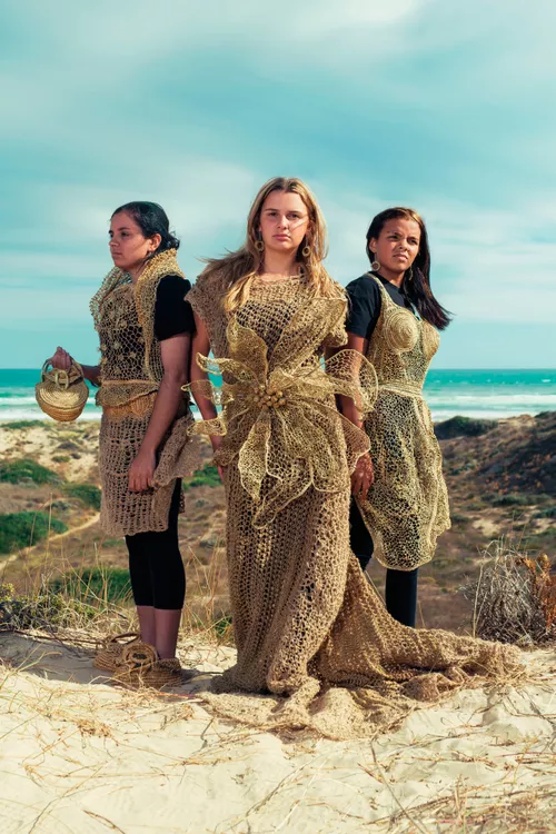 Three women stand in a coastal landscape wearing woven pieces made by First Nations artist Aunty Ellen Trevorrow