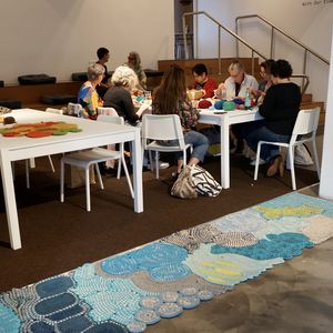 Lynn's large crocheted floor runner in blues and greens with makers crocheting at a table in the background