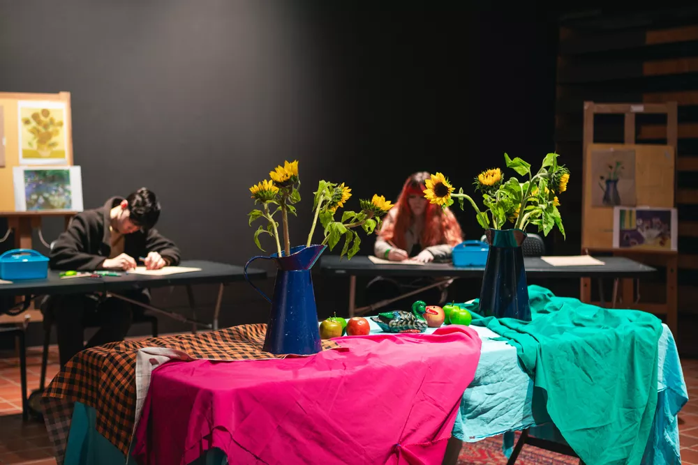 Still life set up in the middle of the room, with fabric draped over the table, sunflowers in jugs, apples and ceramic ducks. Two young people can be seen in the background creating drawings from the scene.