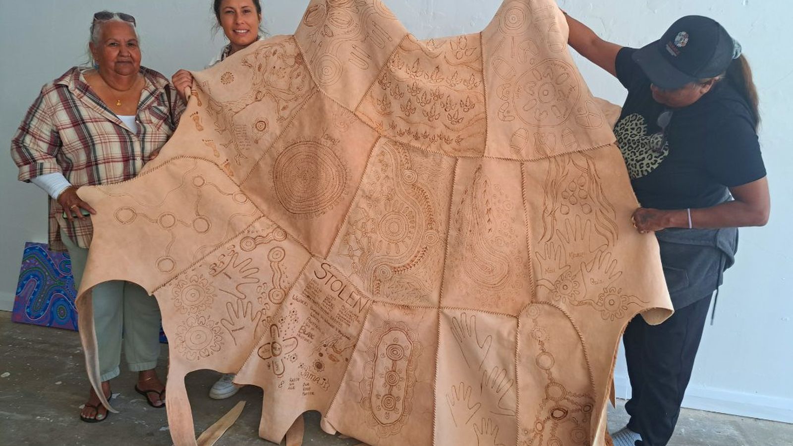 Three women holding up a large cloak made of many pieces of kangaroo skin sewn together with the maker's stories burnt onto the cloak