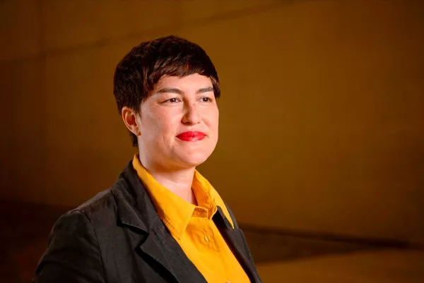 Curator Ellie Buttrose, a young woman with cropped brown hair, smiles while wearing red lipstick, a yellow collared shirt and a black blazer
