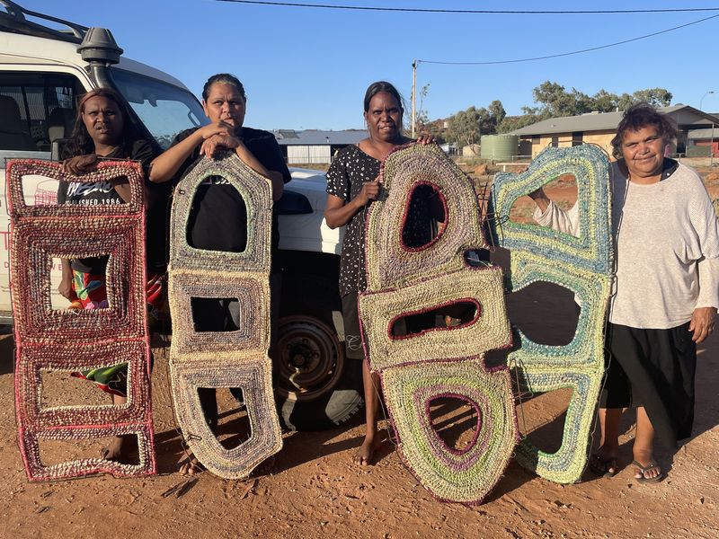 Photo of four women holding up woven sculptures on a red dirt road in Central Australia