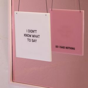 Two square pendants, one white with black text that says " I Didn’t Know What To Say" and the other square is pink with black text, the text says "So I Said Nothing". The squares are hanging on a black cord against a pink mirror surface