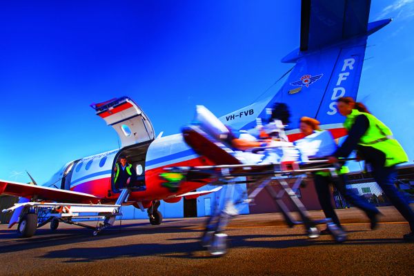 An aircraft with RFDS logo. A patient is lying on a wheeled stretcher, being pushed by a person wearing a high-visibility jacket. 