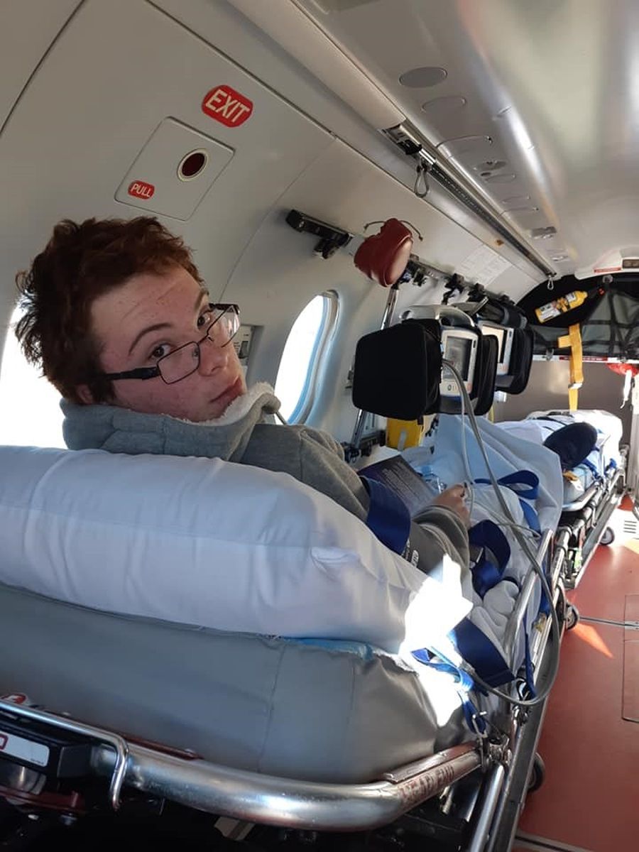 A teenage boy lies on a hospital stretcher in a small aircraft. He is wearing a neck brace and glasses. 