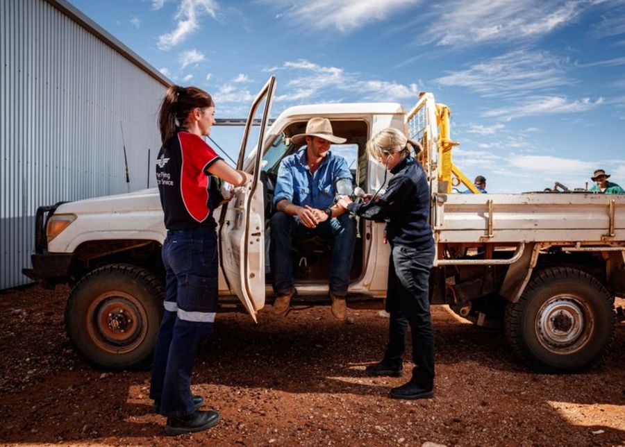 a nurse in an RFDS shirt and a doctor checking the heart rate of a patient who is sitting in a ute