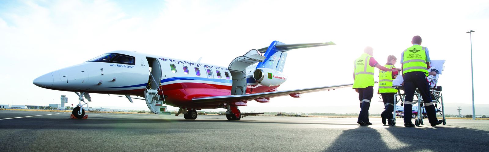 A jet aircraft with an RFDS logo. People in high-vis clothing push a hospital stretcher with a person lying on top. 