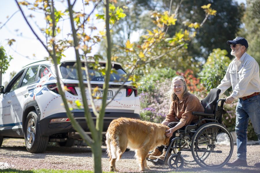 A smiling woman in a wheelchair pats a golden retriever while a volunteer pushes her chair, with an RFDS Community Transport vehicle.