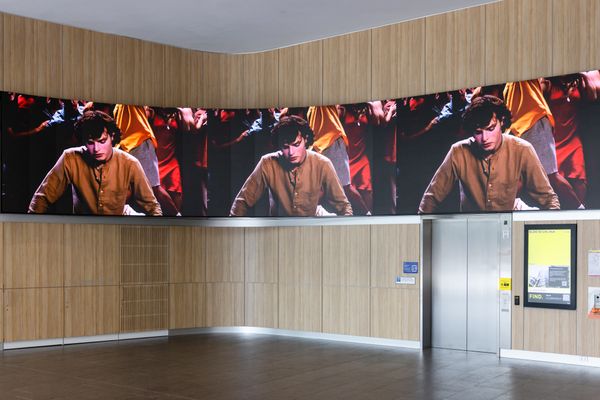 A photo of a large led screen with an image of a young man looking at his reflection in a pool of water. He wears an orange shirt.