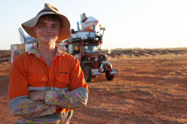 A young man stands in front of an orange tractor. He is a wearing bright orange work shirt and old hat and is smiling at the camera.