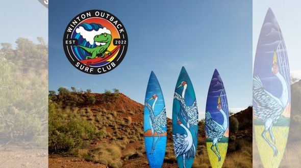 3 surfboards with paintings of birds are pictured standing up in the outback. The dirt is bright orange and sky a rich blue.
