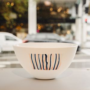 A decorative white bowl with blue, orange and light blue vertical stripes, positioned on a plinth by a window overlooking a busy street.