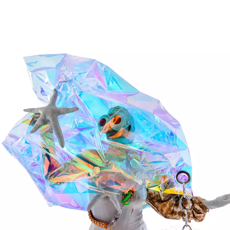 A mid-shot of a performer from the waist up is pictured. The performer is wearing a silver-white sequinned bodysuit and pair of large goggles, made of the same silver material as the bodysuit. On one arm, the performer wears a structured sleeve, made in a tan/brown patterned material. A crystal-like formation, iridescent in colour and somewhat see-through, rests above the performer's head in a peak and flares out slightly to the waist of the performer.