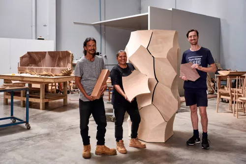 Three people stand smiling with a large-scaled furniture art piece, each holding different wooden elements.