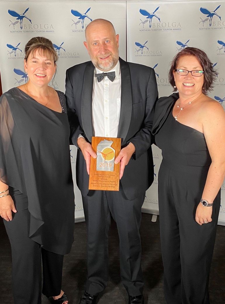 Three people in black formal wear smile at the camera. They are holding a trophy that reads 2019 BROLGA AWARD. 
