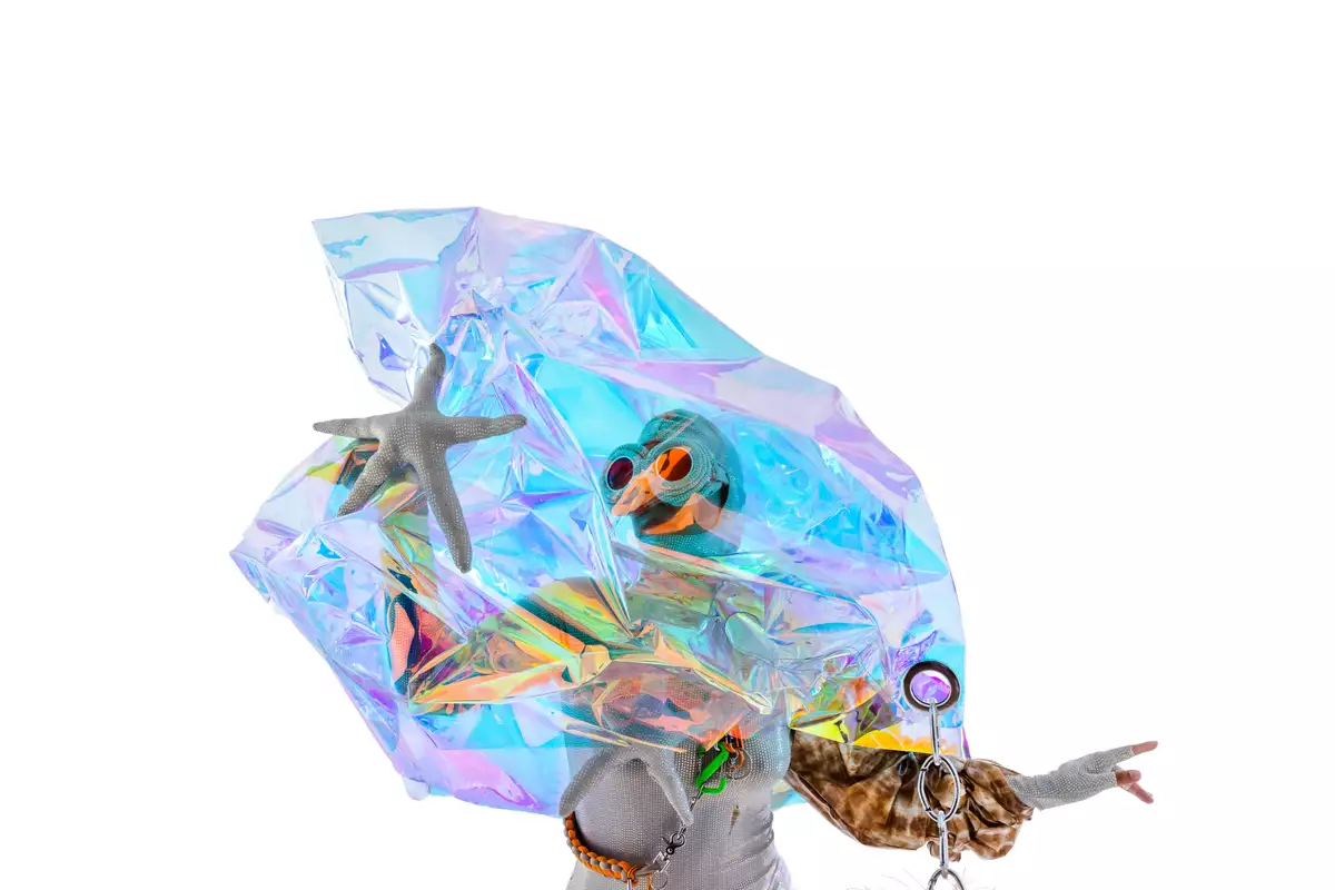 A mid-shot of a performer from the waist up is pictured. The performer is wearing a silver-white sequinned bodysuit and pair of large goggles, made of the same silver material as the bodysuit. On one arm, the performer wears a structured sleeve, made in a tan/brown patterned material. A crystal-like formation, iridescent in colour and somewhat see-through, rests above the performer's head in a peak and flares out slightly to the waist of the performer.