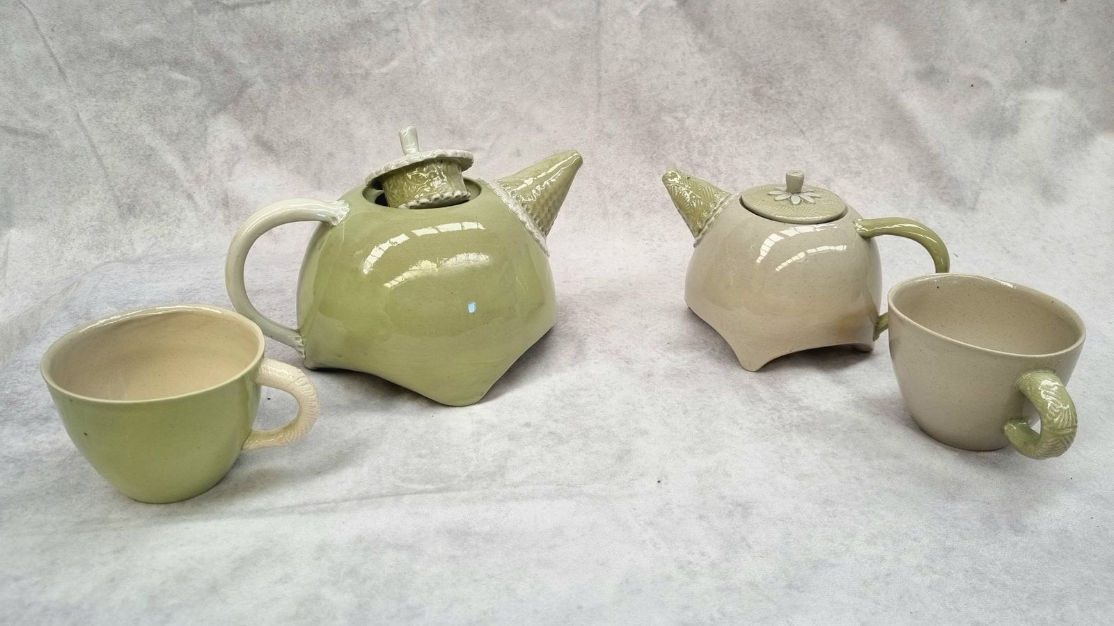 Light green & cream ceramic teapots & matching cups against a grey cloth background