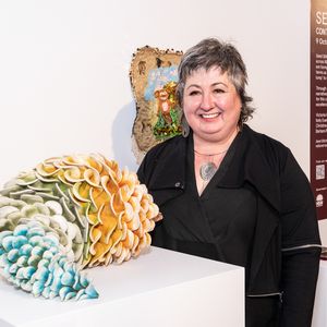 Women with short grey hair smiling at camera next to textile sculpture on white plinth.