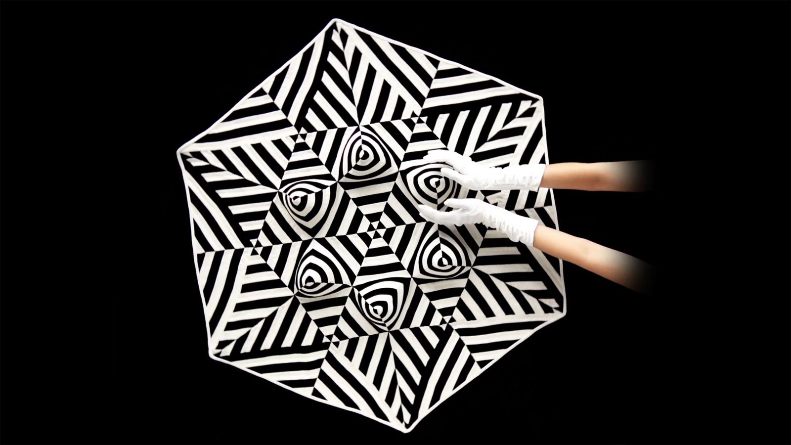 two arms and white gloved hands cupping part of the pattern created by the black and white illusion quilt in the background