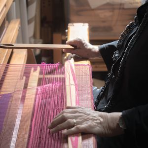 Photo of two hands holding a wooden shaft in front of pink strings of a large weaving loom.