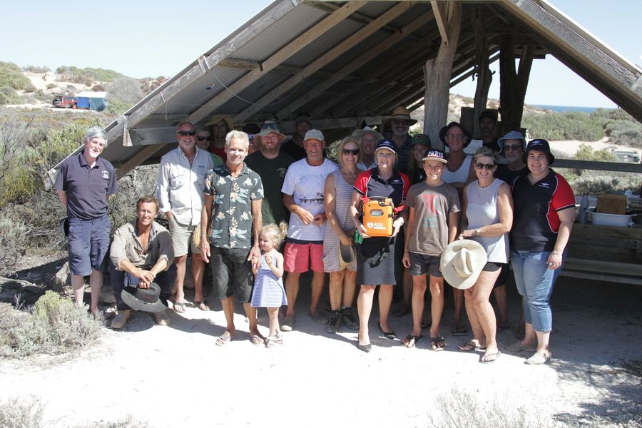 5 people stand under a basic wooden hut at a beach. They all smile at the camera. 