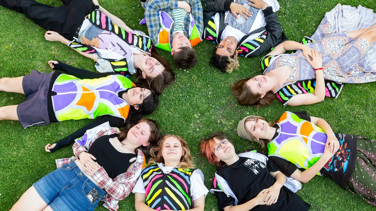 A group of young people laying on the grass in a circle with their heads close together.