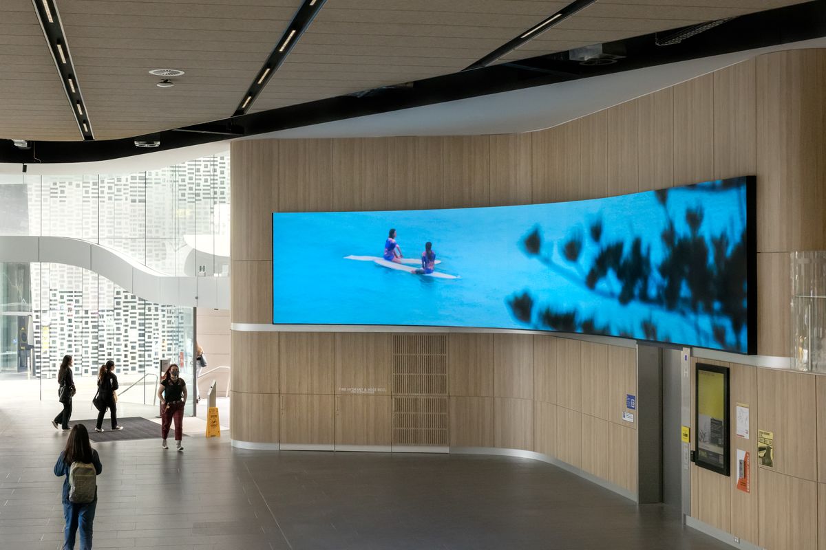 A large digital screen in a public space depicting two surfers floating in the waves