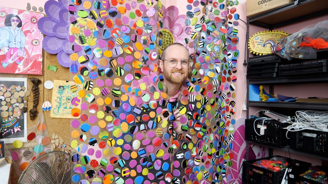 An image of the artist poking his head through his colourful curtains made of hand painted cardboard pieces on tulle, with the background of his studio