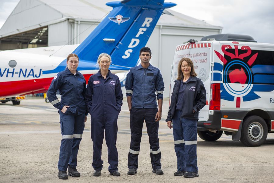 Four Royal Flying Doctor Service crew members in navy flight suits standing on the tarmac in front of an RFDS aircraft and patient transport vehicle.
