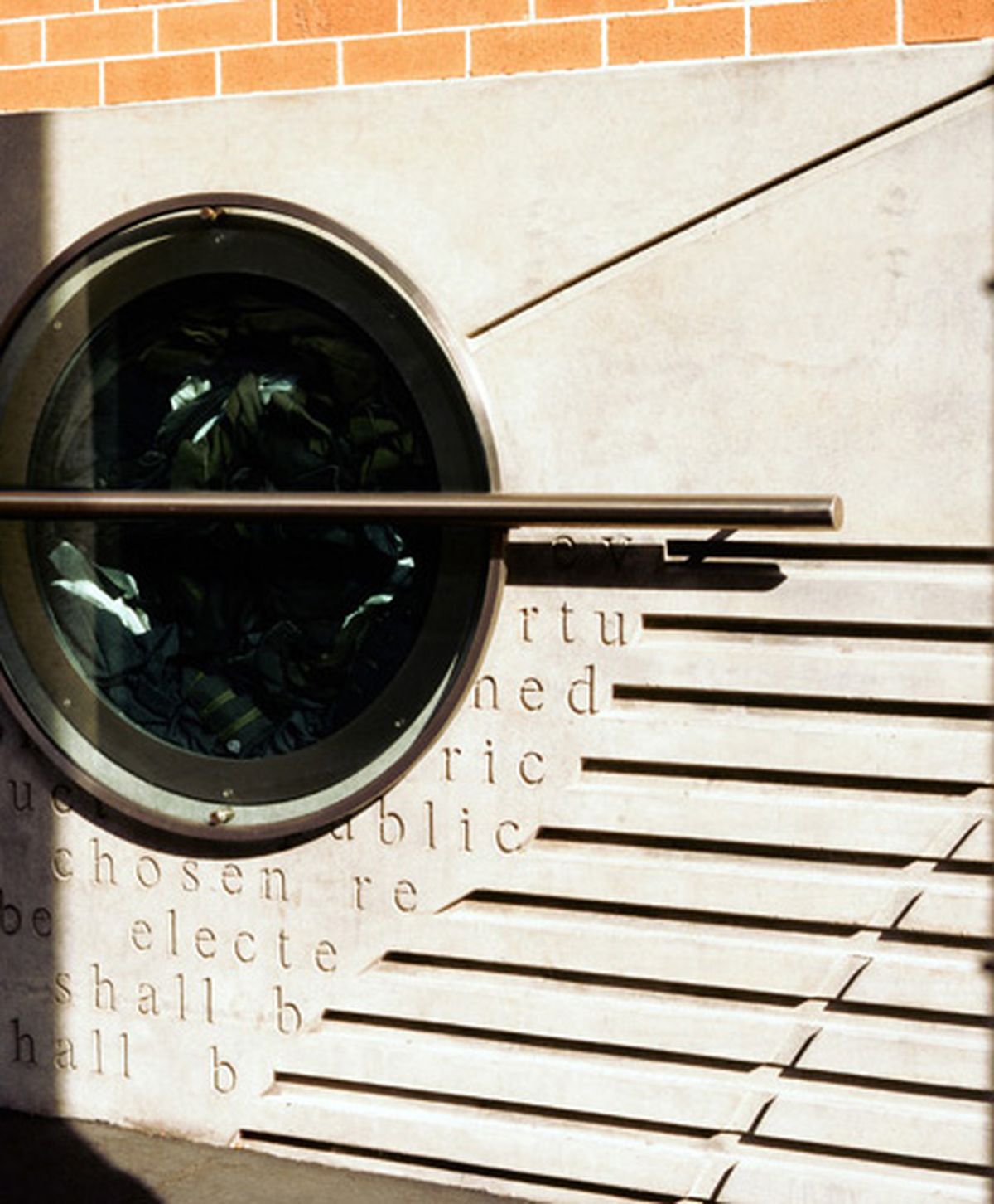 Photo of a concrete wall with horizontal indents in it and a circular window inset to the left of the image with clothing inside.