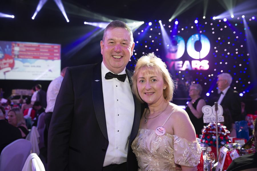 A man and woman in formal clothes smile at the camera. They appear to be at a gala event. 
