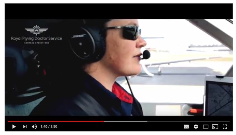 A woman wearing a red and navy blue RFDS shirt and cargo pants. She is standing next to a RFDS aircraft and is smiling. 