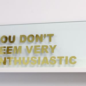 A glass panel with the text "YOU DON'T SEEM VERY ENTHUSIASTIC" written in gold capital letters, placed on a white shelf against a white wall.