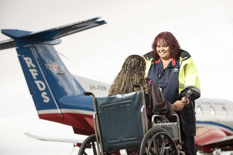 A lady is sat in a wheelchair. She holds hands with a lady in RFDS uniform, they are smiling. A RFDS aircraft is in the background. 