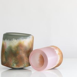Two vases, one green and another pink. Both placed on a white surface, showcasing distinct hues of green, orange, green and white. The pink vase in laying horizontally on the surface